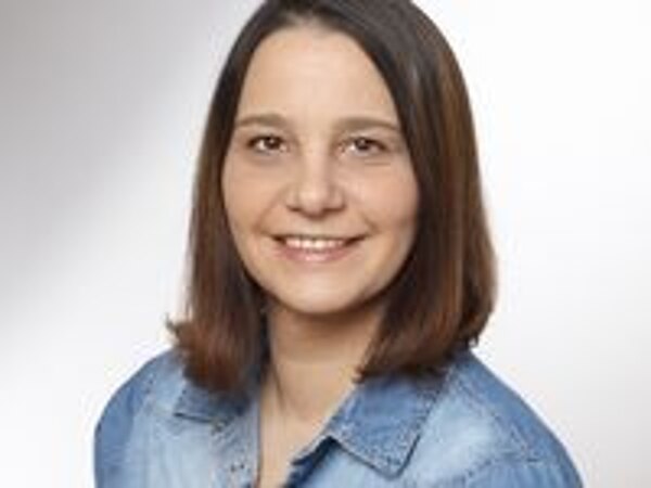 Ein Porträt einer Frau mit mittellangen, braunen Haaren. Sie trägt ein denimfarbenes Hemd und lächelt freundlich in die Kamera. Der Hintergrund ist hell und neutral gestaltet.