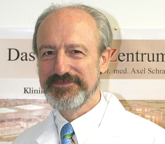 Porträt eines älteren Mannes mit grauem Bart in weißem Arztkittel und Krawatte. Er lächelt leicht und schaut in die Kamera; Hintergrund mit unscharfem Schild, nebensächlich.