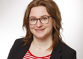 Theresa Scheurer Porträtfoto einer lächelnden Frau mit schulterlangen braunen Haaren und Brille, in schwarzem Blazer über rot-weiß gestreiftem Oberteil, vor hellem neutralem Hintergrund.