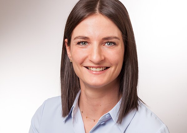 Porträtfoto einer lächelnden Frau mit schulterlangem, glattem, dunkelbraunem Haar, leichter Goldkette und hellblauer Bluse. Sie blickt direkt in die Kamera vor neutralem, hellem Hintergrund.