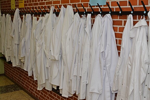 An einer Wand hängen mehrere weiße Kittel auf einem Kleiderständer. Die Kittel sind ordentlich nebeneinander aufgehängt. Der Hintergrund besteht aus einer roten Ziegelmauer. Die Szene vermittelt eine typische Umgebung, in der Labor- oder medizinische Arbeiten stattfinden.