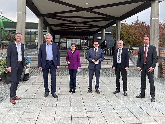 Sechs Erwachsene in formeller Kleidung stehen mit Abstand vor dem Eingang eines modernen Gebäudes und blicken zur Kamera. Sie wirken wie zu einem offiziellen Termin versammelt.