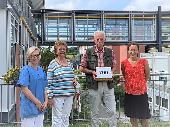 Vier Erwachsene stehen nebeneinander vor einem modernen Gebäude. Der Mann in der Mitte hält ein Schild mit der Zahl 700. Gruppenfoto, das einen erreichten Meilenstein oder eine Übergabe markiert.