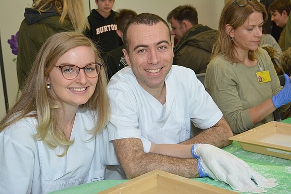 Zwei lächelnde Personen in weißen Kitteln sitzen an einem Tisch mit einem grünen Tuch. Vor ihnen befinden sich flache Schalen. Im Hintergrund sind weitere Personen zu sehen, die ebenfalls an der Aktivität teilnehmen.