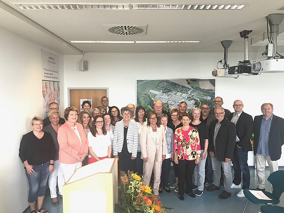 Gruppe von etwa 25 Erwachsenen steht in einem hellen Konferenzraum und blickt in die Kamera. Im Vordergrund ein Rednerpult mit Blumen. Die Szene wirkt wie ein offizielles Treffen, Gruppenfoto oder eine Ehrung.