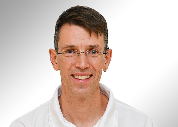 Porträt eines lächelnden erwachsenen Mannes mit kurzem braunen Haar, schmaler Brille und kleinem Ohrring, in weißem Poloshirt vor hellem, neutralem Hintergrund.