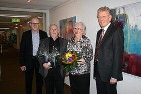 Vier Personen stehen in einem Flur, einige halten Blumen und Geschenke. Sie scheinen einen feierlichen Moment zu teilen, möglicherweise eine besondere Gelegenheit oder Verabschiedung. Die Wände sind mit abstrakter Kunst dekoriert.
