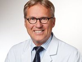 Dr. med Paul-Wilhelm Zühlke Ein lächelnder Arzt in weißem Kittel mit einer hellblauen Hemd und einer dunklen Krawatte. Er trägt runde Brille und hat kurzes, blondes Haar. Der Hintergrund ist hell und schlicht, um den Fokus auf den Arzt zu legen.