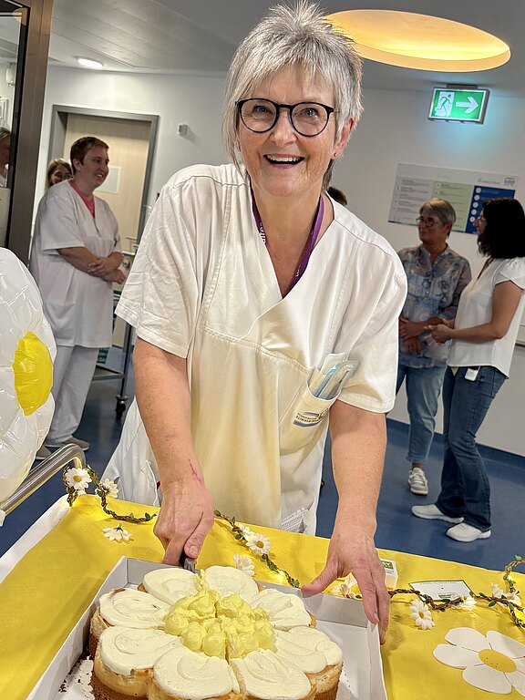 Ältere Frau in weißer Klinikbekleidung lächelt und präsentiert einen in Blütenform dekorierten Kuchen auf einem gelb geschmückten Tisch. Im Hintergrund stehen Kolleginnen und Kollegen auf einem Krankenhausflur. Die Szene wirkt wie eine kleine Feier oder Pause im Team.