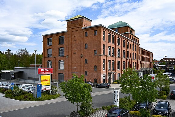 Großes historisches Backsteingebäude mit mehreren Stockwerken, vielen hohen Fenstern und zwei Dachaufbauten. Davor Parkplätze mit wenigen Autos und etwas Grün. Tageslicht, teils bewölkter Himmel. Wirkt wie eine ehemals industrielle Anlage, heute gewerblich oder büroartig genutzt.