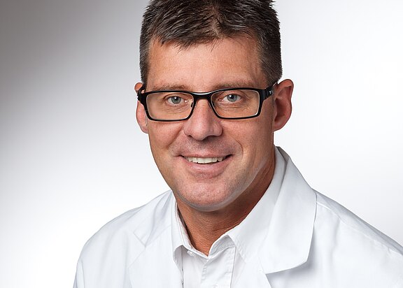 Porträt einer lächelnden Person mit kurzer Frisur und schwarzer Brille, im weißen Kittel vor neutralem Hintergrund.