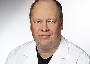 Porträt eines mittelalten Mannes in weißem Kittel vor neutralem Hintergrund, der direkt in die Kamera blickt, mit neutralem Ausdruck.