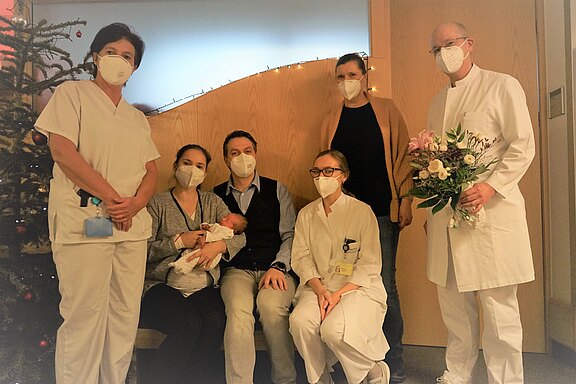 Gruppenfoto im Krankenhaus: Eine Mutter hält ihr Neugeborenes, der Vater sitzt daneben. Mehrere Pflegekräfte und Ärztinnen/Ärzte mit Masken stehen bzw. knien daneben; eine Person hält einen Blumenstrauß. Szene nach einer Geburt.