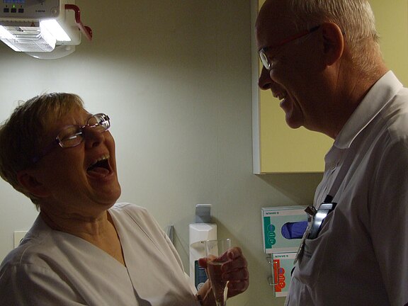 Zwei medizinische Fachkräfte in weißen Uniformen stehen einander gegenüber und lachen herzlich in einem Klinikraum. Eine Person hält ein Glas. Die Szene vermittelt Kollegialität und Freude.