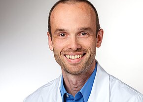 Dr. med. Michael Kryk Porträt eines lächelnden Mannes in weißem Arztkittel und blauem Hemd vor neutralem Hintergrund; er schaut direkt in die Kamera. Das Bild vermittelt den Eindruck eines freundlichen medizinischen Fachpersonals.