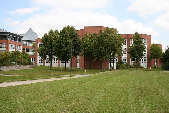 Modernes Hochschulgebäude aus rotem Klinker mit vielen Fenstern, umgeben von Rasen und Bäumen; ein Weg führt durch die Grünfläche unter blauem Himmel.