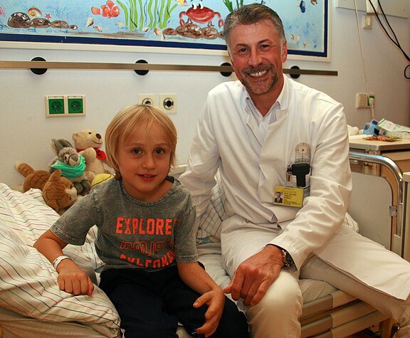 Ein lächelnder Arzt im weißen Kittel sitzt neben einem Jungen auf einem Krankenhausbett. Der Junge trägt ein T-Shirt, wirkt ruhig und hat ein Armband am Handgelenk. Beide schauen in die Kamera; die Szene vermittelt freundliche, vertrauensvolle Betreuung in einer Kinderklinik.