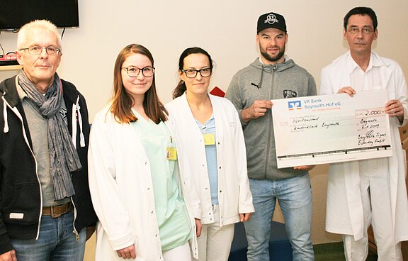 Fünf Personen stehen in einem Krankenhausraum. Drei tragen weiße Kittel. Eine Person in Freizeitkleidung hält einen großen symbolischen Scheck; daneben ein Arzt. Szene einer Spendenübergabe an medizinisches Personal.