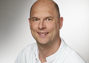 Porträtfoto: Ein lächelnder, glatzköpfiger Mann in weißem Kittel blickt direkt in die Kamera. Neutraler Hintergrund, sachlich-professionelle Darstellung.