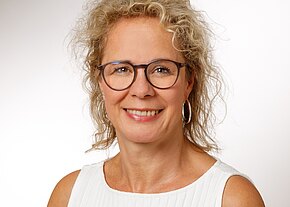 Porträtfoto einer lächelnden Frau mit lockigem blondem Haar und runder Brille. Sie trägt ein ärmelloses weißes Oberteil und runde Ohrringe. Heller, neutraler Hintergrund; Fokus auf ihrem freundlichen Gesicht.