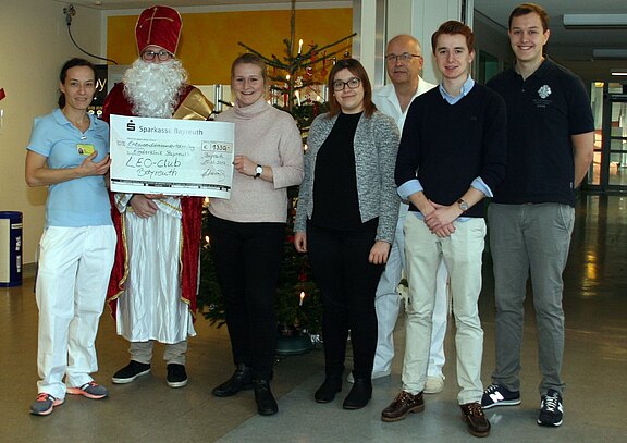 Gruppe in Klinikflur, neben Weihnachtsbaum. Eine Person als Nikolaus hält mit anderen einen übergroßen Spendenscheck der Sparkasse Bayreuth. Der Scheck ist für den LEO-Club Bayreuth. Alle lächeln – symbolische Spendenübergabe zur Weihnachtszeit.
