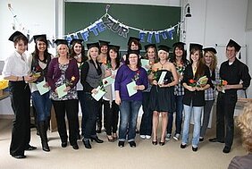 Eine Gruppe von Absolventen steht zusammen in einem Klassenzimmer. Sie tragen schwarze Graduationshüte und halten Dokumente sowie Blumen in den Händen. Im Hintergrund hängt ein Banner mit der Aufschrift „HERZLICHEN GLÜCKWUNSCH“. Die Atmosphäre ist festlich und feierlich.