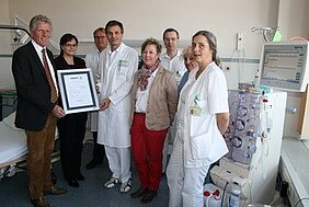 Eine Gruppe von sieben Personen in Krankenhausuniformen und Zivilkleidung steht zusammen in einem Raum. Sie halten ein Zertifikat in der Mitte der Gruppe, das eine Auszeichnung verkündet. Im Hintergrund ist ein medizinisches Gerät sichtbar.