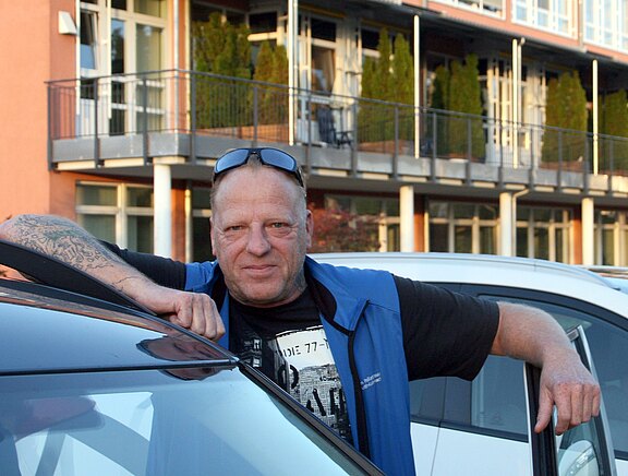 Ein Mann lehnt entspannt an der geöffneten Autotür und blickt in die Kamera. Er trägt eine blaue Weste, ein schwarzes T‑Shirt und eine Sonnenbrille auf dem Kopf.