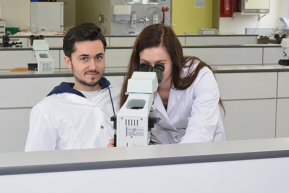 Zwei Personen in weißen Laborkitteln arbeiten in einem Labor. Eine Person schaut durch ein Mikroskop, während die andere sie von der Seite beobachtet. Im Hintergrund sind weitere Labormöglichkeiten sichtbar.