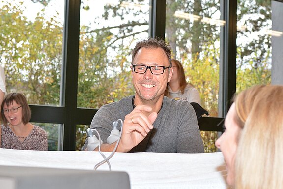 Ein lächelnder Mann mit Brille sitzt an einem Tisch, während er mit einer anderen Person spricht. Im Hintergrund sind weitere Personen zu sehen, die lachen. Die Szene vermittelt eine freundliche und gesellige Atmosphäre in einem hellen Raum mit Fenstern und Bäumen im Hintergrund.