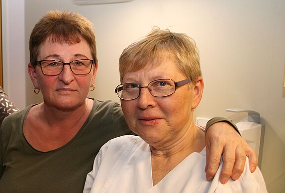 Zwei ältere Frauen mit Brille sitzen dicht beieinander; eine legt der anderen den Arm um die Schulter. Beide blicken freundlich in die Kamera in einem Innenraum.