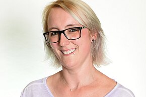 Porträt einer lächelnden Person mit kurzem blondem Haar, schwarzen rechteckigen Brillen, kleinem Lippenpiercing und Ohrstecker. Sie trägt ein helles T‑Shirt und eine feine Halskette. Neutraler, heller Hintergrund.