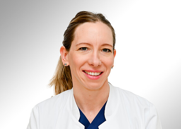 Porträt einer lächelnden medizinischen Fachkraft in weißem Kittel und dunkelblauem Oberteil, vor hellem Hintergrund. Haare zum Zopf gebunden, Blick in die Kamera.