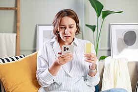 Eine besorgte Frau in einem gestreiften Hemd sitzt auf einem Sofa. Sie hält ein Telefon in der einen Hand und eine gelbe Karte in der anderen. Ihr Gesichtsausdruck zeigt Besorgnis, während sie auf den Bildschirm schaut.