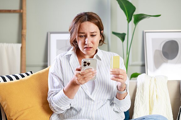 Eine besorgte Frau in einem gestreiften Hemd sitzt auf einem Sofa. Sie hält ein Telefon in der einen Hand und eine gelbe Karte in der anderen. Ihr Gesichtsausdruck zeigt Besorgnis, während sie auf den Bildschirm schaut.