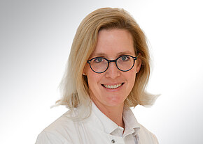 Dr. med. univ. Sophie Lau Porträt einer lächelnden Person mit schulterlangem blondem Haar und runder Brille in weißem Arztkittel vor hellem, neutralem Hintergrund. Kopf und Oberkörper leicht schräg zur Kamera, freundlicher, professioneller Ausdruck.