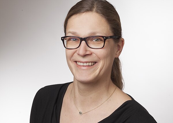 Porträt einer lächelnden Frau mit Brille, zurückgebundenen braunen Haaren und schwarzem Oberteil. Sie trägt eine feine Kette. Heller, neutraler Hintergrund, Kopf und Schultern im Bild.