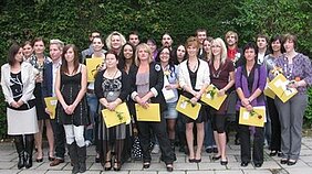 Eine Gruppe von etwa 20 Personen steht zusammen im Freien, lächelt und hält Urkunden in den Händen. Die Anwesenden sind frauen- und männergemischt, in verschiedenen Kleidungsstilen, und scheinen an einer Feier oder Zeremonie teilzunehmen. Im Hintergrund sind Pflanzen zu sehen.
