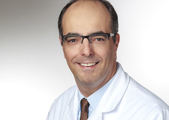 Porträt eines lächelnden Mannes mit Brille in weißem Arztkittel und Krawatte vor neutralem Hintergrund. Nahaufnahme bis zur Brust, professionelles Foto, vermittelt Vertrauenswürdigkeit und medizinischen Kontext.