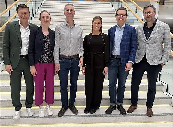 Sechs Erwachsene posieren lächelnd auf einer breiten Treppe in einem modernen Gebäude. Sie stehen nebeneinander, tragen überwiegend Business-Casual-Kleidung in unterschiedlichen Farben. Geländer links und rechts, helles Treppenhaus. Gruppenfoto, frontale Ansicht, entspannte Stimmung.