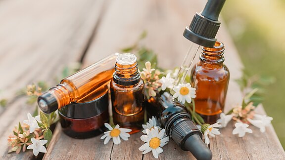 Mehrere braune Tropfflaschen mit ätherischen Ölen, teils offen mit Pipette, umgeben von weißen Blüten auf Holz. Darstellung natürlicher Aromatherapie- oder Pflegeprodukte.