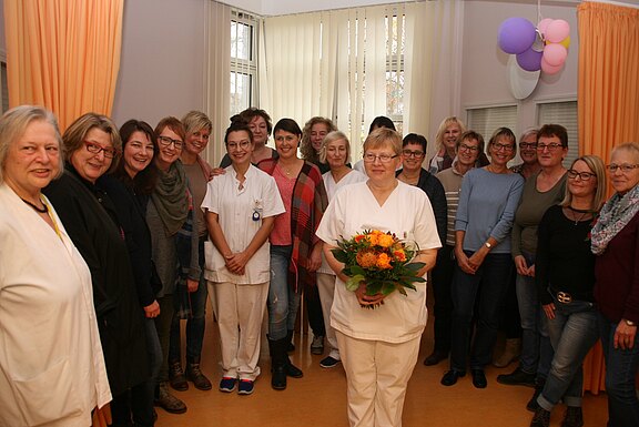 In einem Klinikraum steht eine Pflegekraft mit Blumenstrauß im Vordergrund. Hinter ihr posiert eine größere Gruppe Kolleginnen und Kollegen, teils in weißer Arbeitskleidung. Freundliche, gemeinschaftliche Atmosphäre.
