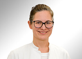 Porträt einer lächelnden Frau mit dunkler Brille und zusammengebundenen Haaren in weißem Kittel vor hellem, neutralem Hintergrund. Kopf und Oberkörper im Vordergrund.