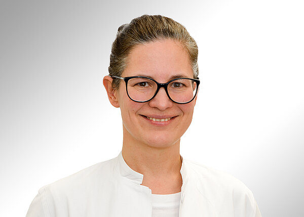 Porträt einer lächelnden Frau mit dunkler Brille und zusammengebundenen Haaren in weißem Kittel vor hellem, neutralem Hintergrund. Kopf und Oberkörper im Vordergrund.