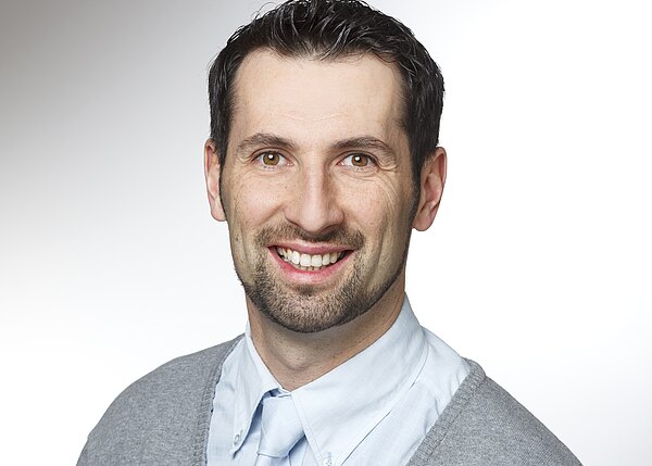 Porträtfoto: Ein lächelnder erwachsener Mann mit kurzen dunklen Haaren und kurzem Bart. Er trägt ein hellblaues Hemd und eine graue Strickjacke. Neutraler, heller Hintergrund. Fokus auf freundlichem, professionellem Ausdruck.