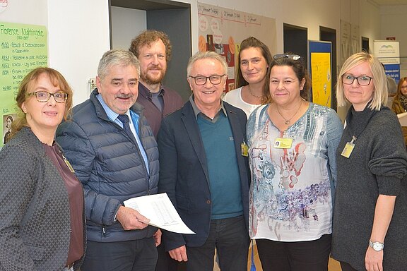 Eine Gruppe von sechs Personen steht zusammen in einem Raum. Sie lächeln und tragen Namensschilder. Im Hintergrund sind verschiedene Informationsplakate sichtbar. Ein Mann hält ein Dokument in der Hand. Die Atmosphäre wirkt freundlich und einladend.