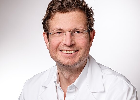 Porträtfoto: Ein lächelnder Mann mit Brille trägt einen weißen Kittel und schaut direkt in die Kamera. Neutraler, hellgrauer Hintergrund.