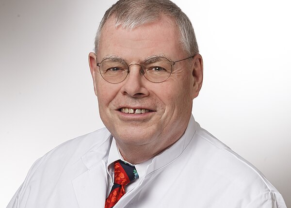 Porträt eines älteren Mannes mit Brille in weißem Kittel und roter Krawatte. Er lächelt freundlich, schaut in die Kamera; neutraler Hintergrund.