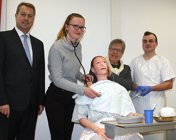 In einem Schulungsraum übt eine Pflegeschülerin mit Stethoskop an einer realistischen Patientensimulationspuppe. Neben ihr stehen eine Ausbilderin, ein weiterer Pflegeschüler in Schutzkleidung und eine Person im Anzug. Das Bild vermittelt Pflegeausbildung und praktische Übung am Simulator.