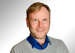 Porträtfoto: Ein lächelnder erwachsener Mann mit hellem Haar, trägt blaues Hemd unter grauem Pullover, schaut direkt in die Kamera vor neutralem, hellem Hintergrund.